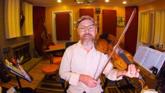 Casey Driessen in Otherlands, sitting in his studio, holding his fiddle and bow, and smiling.
