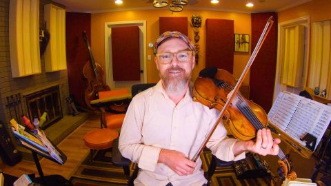 Casey Driessen in Otherlands, sitting in his studio, holding his fiddle and bow, and smiling.