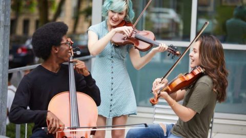 Three students from the Berklee Global Strings program play their string instruments out on the street, one cellist and two violinists. They are looking at each other and smiling.