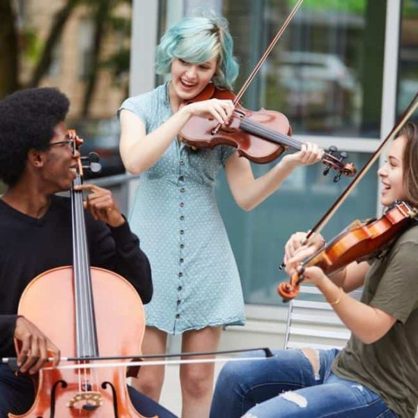 Three students from the Berklee Global Strings program play their string instruments out on the street, one cellist and two violinists. They are looking at each other and smiling.