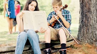 5 Great Summer Music Camps in New England for String Players. The image shows two kids at Kinhaven Music School sitting by the base of a large tree, a girl who holds up sheet music for a boy who look at the music intently while holding his violin in the guitar position.